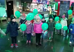 Widok na dzieci trzymające w rękach zielone baloniki. W tle widać bar, plakaty i afisze kinowe.